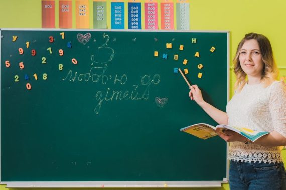 A female teacher pointing to a chalkboard with numbers in a colorful classroom.